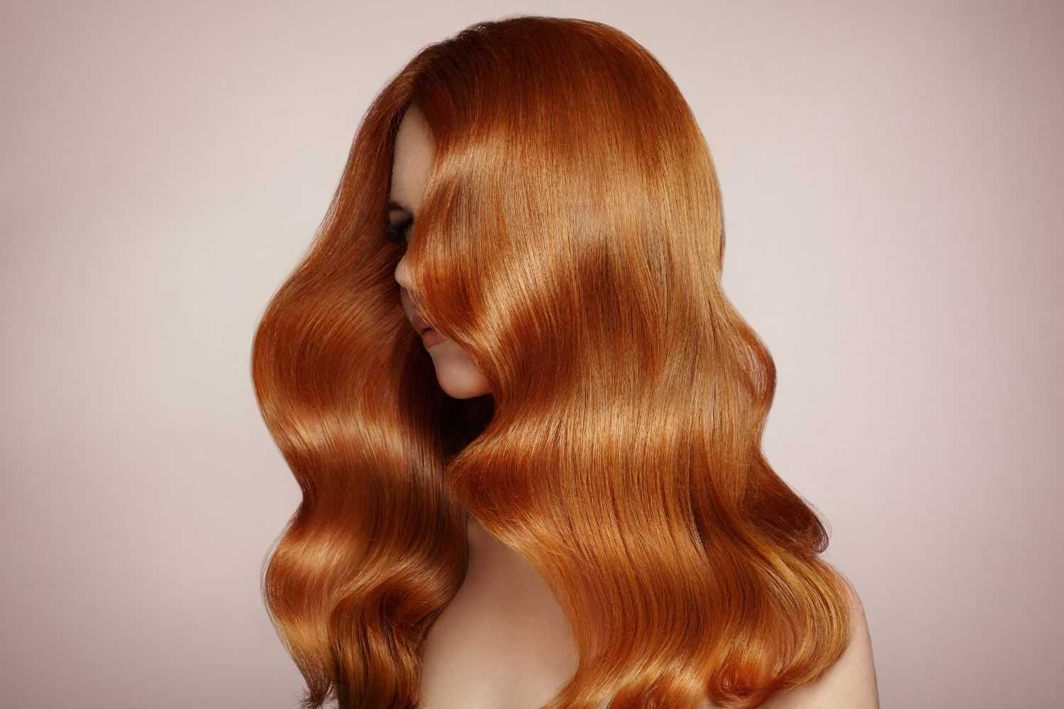 Woman with long, wavy, shiny red hair covering part of her face against a plain background.